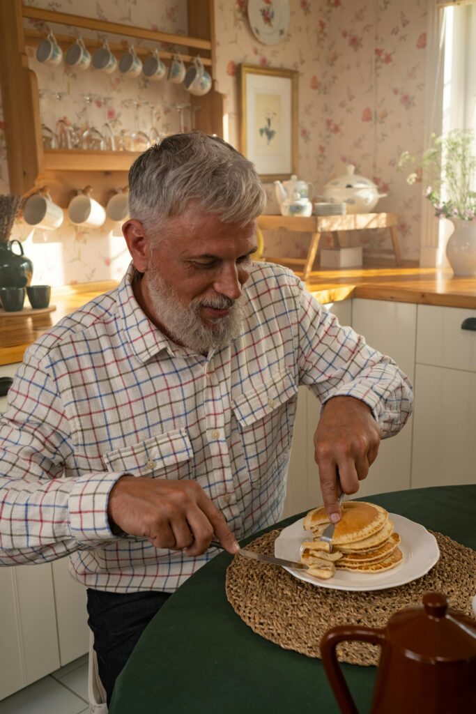 An elderly man with a beard enjoys a stack of pancakes at a cozy kitchen table.